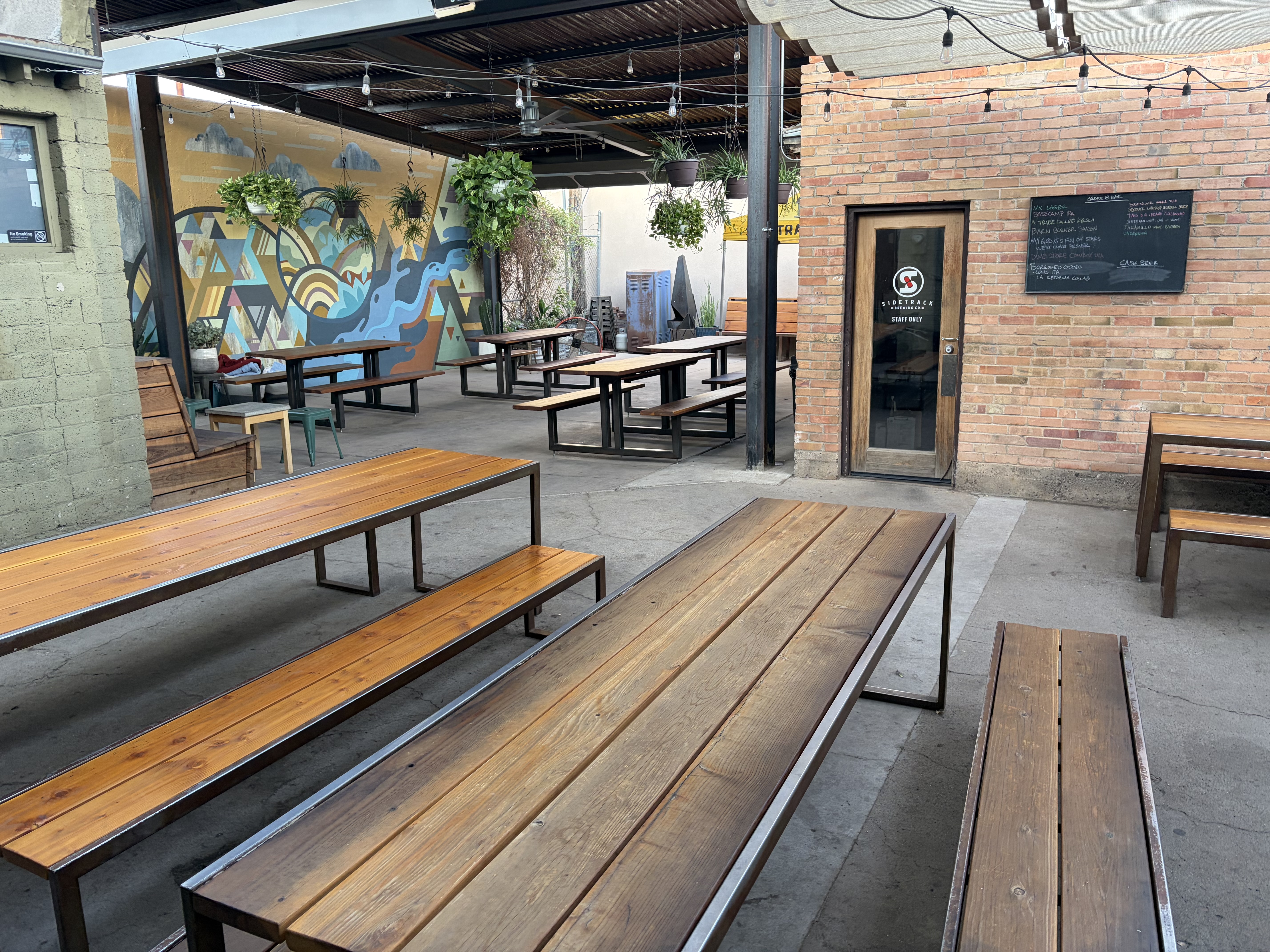 Outdoor patio with picnic tables, string lights overhead, hanging plants, and a large mural of stylised mountains and sky.