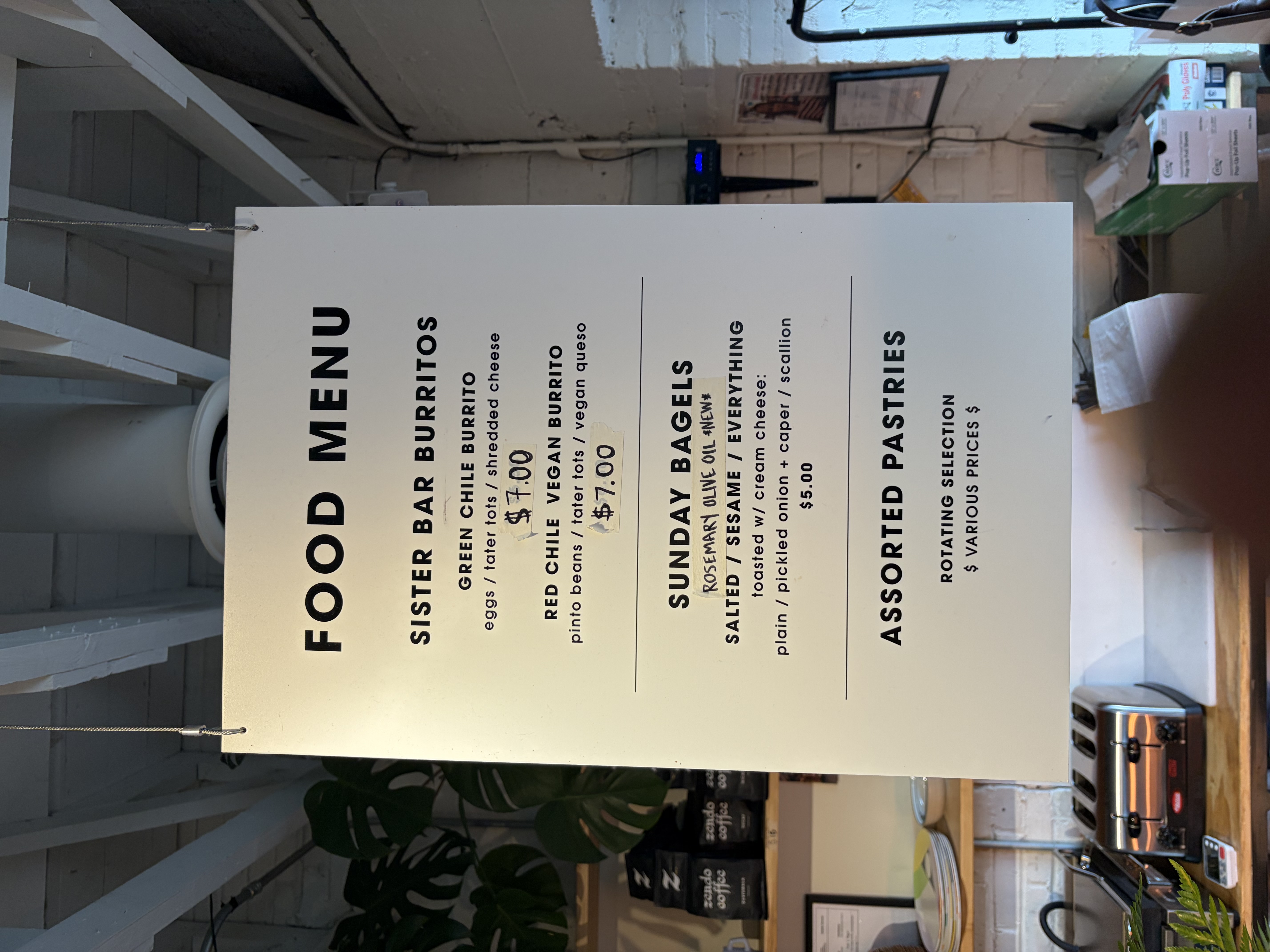 Food menu board: Sister Bar Burritos, green chile burrito with eggs, tater tots and shredded cheese for 7 dollars; red chile vegan burrito 7 dollars; Sunday bagels 5 dollars; assorted pastries.