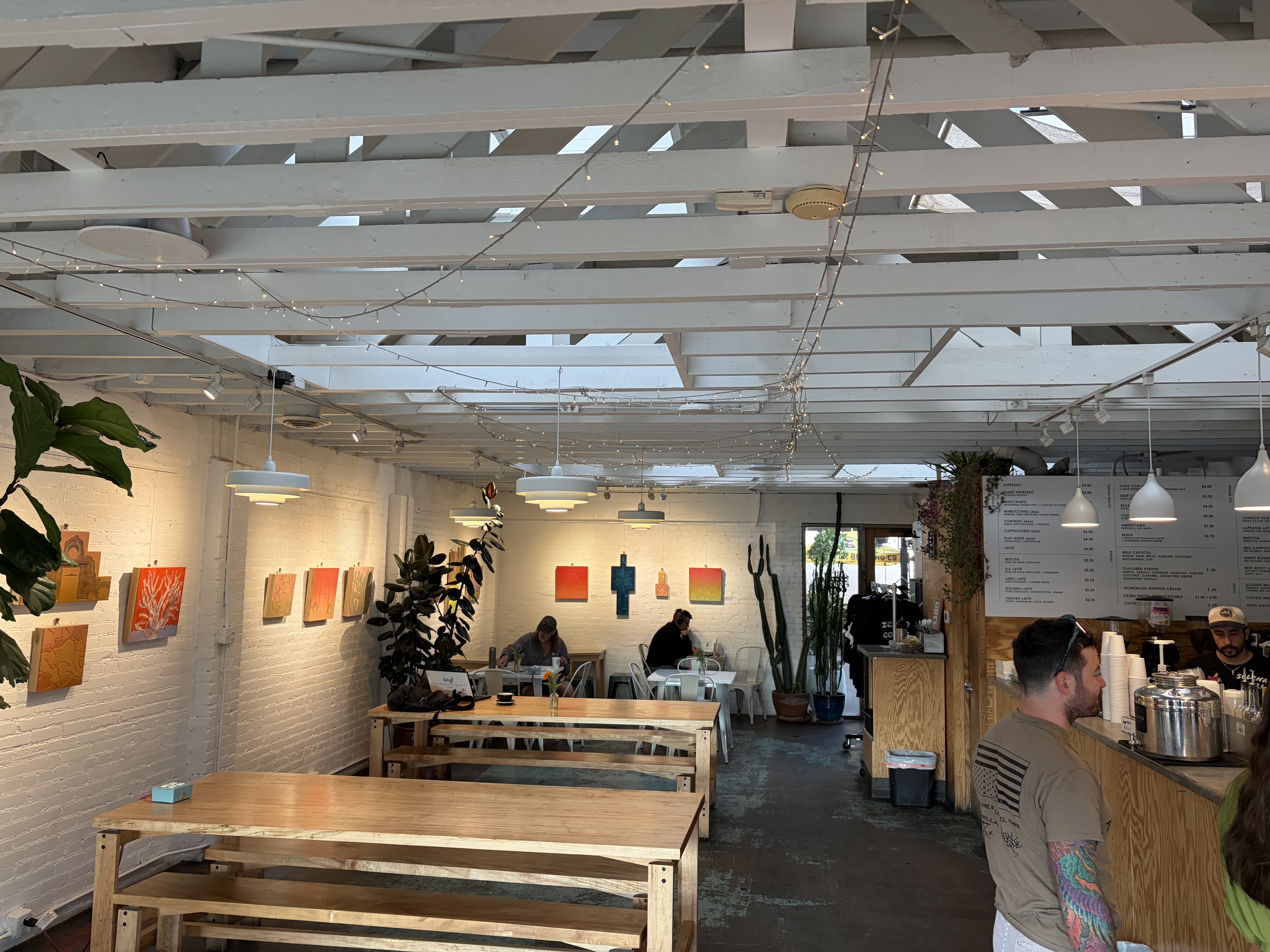 Interior: white painted brick, white exposed roof joists, string lights, pendant lamps, long communal wooden tables, local paintings on the wall, menu boards behind a wooden counter.