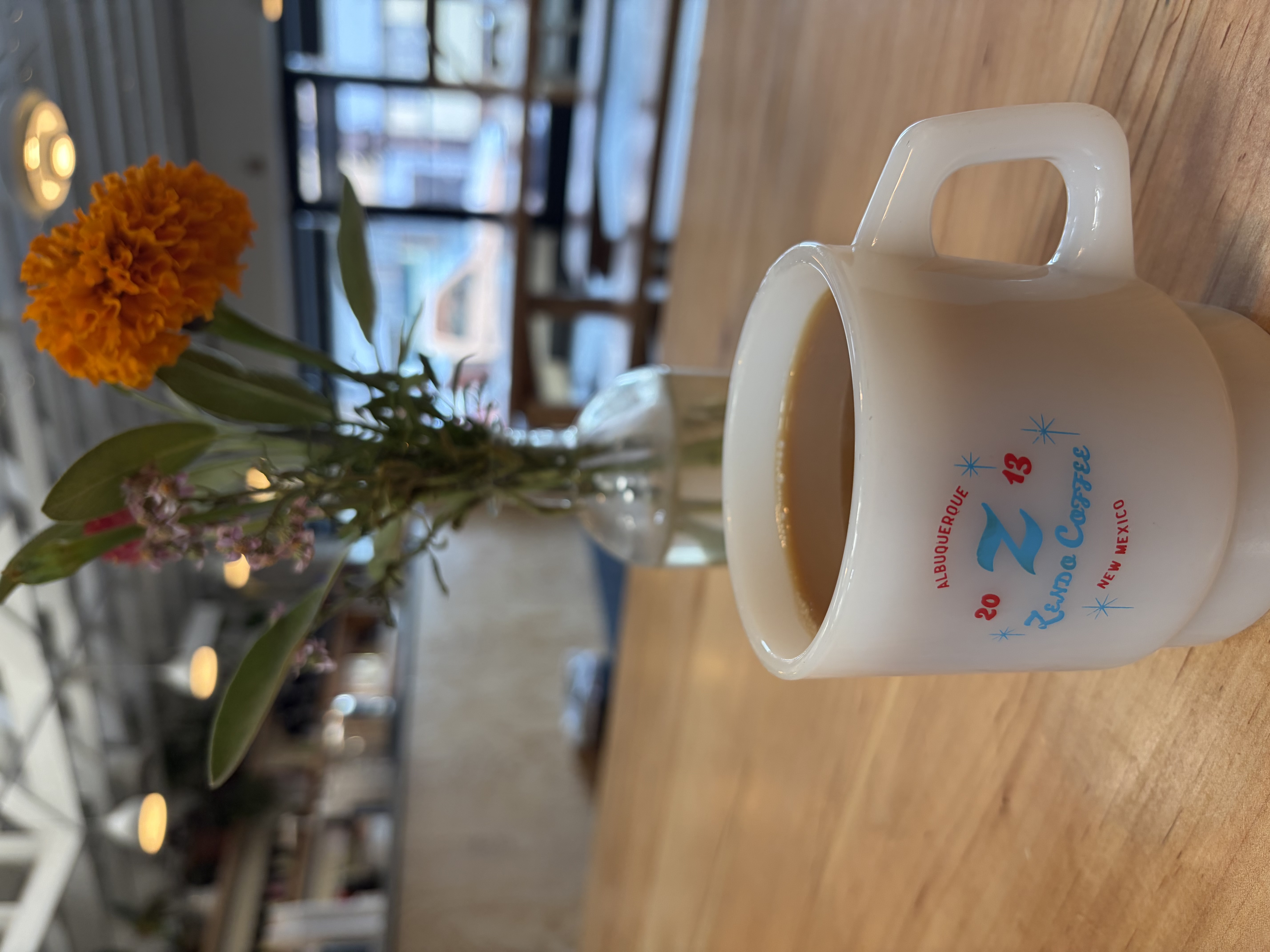 An Americano in a white Zendo Coffee mug printed with a Z, 2013, Albuquerque New Mexico, beside a marigold in a glass vase on a wooden table.